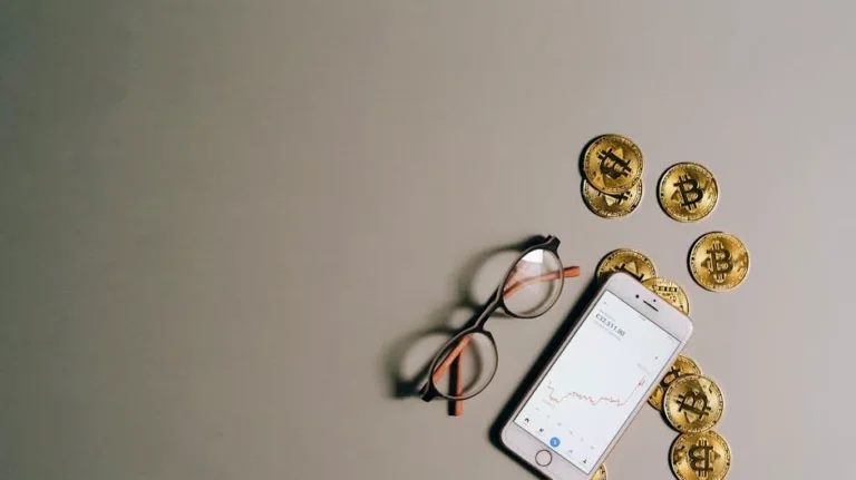 Flat lay of Bitcoin coins, smartphone with graph, and glasses on gray surface.