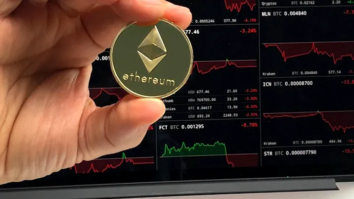 A hand holds an Ethereum coin in front of a laptop displaying cryptocurrency trading charts.