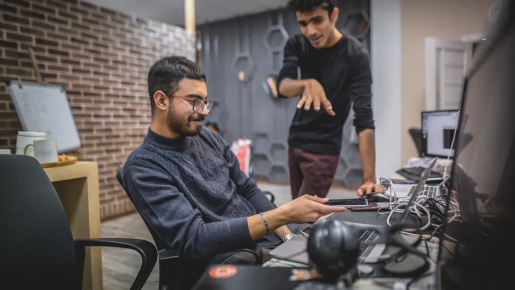 standing man next to sitting man in front of computer in room