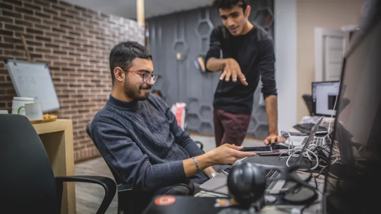 standing man next to sitting man in front of computer in room