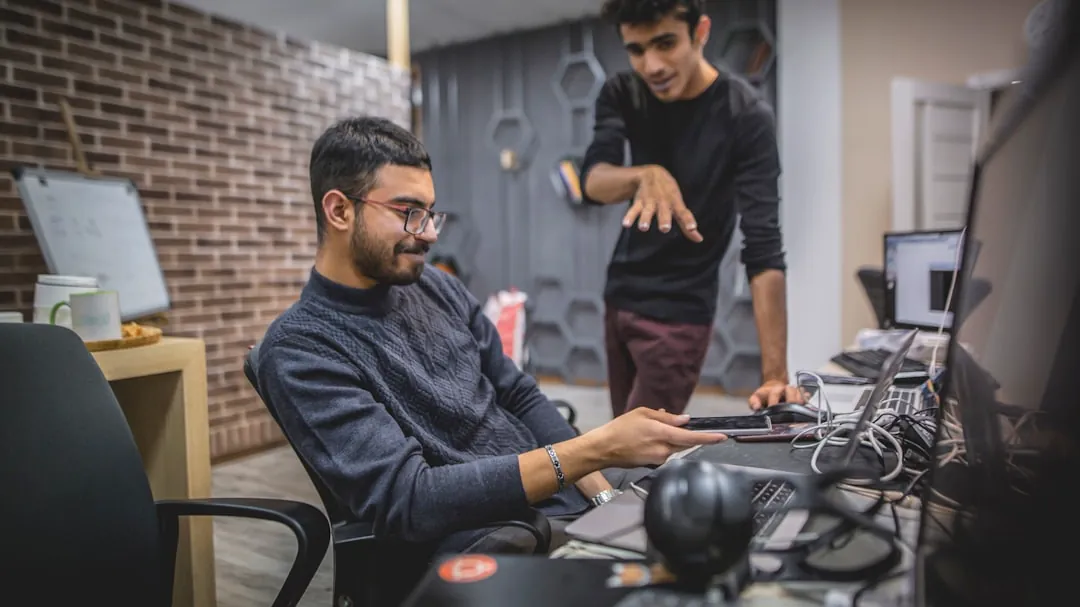 standing man next to sitting man in front of computer in room